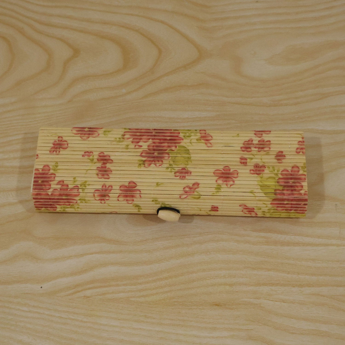 A closed, rectangular bamboo box with a pink floral pattern and roll-top lid, resting on a light wooden surface.
