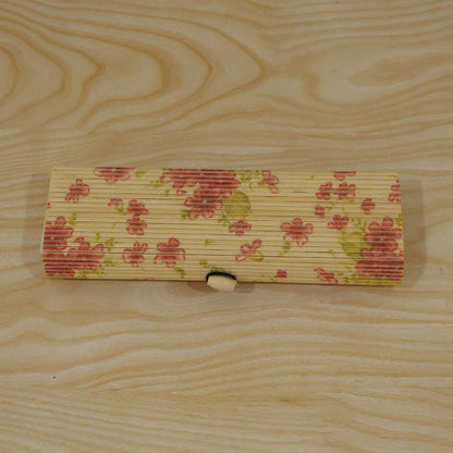 A closed, rectangular bamboo box with a pink floral pattern and roll-top lid, resting on a light wooden surface.