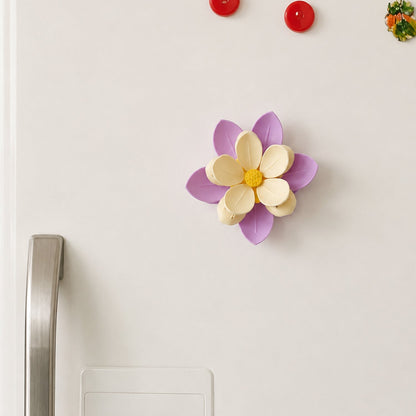 A purple and yellow flower clip magnet attached to a white refrigerator door, with red magnets above it.