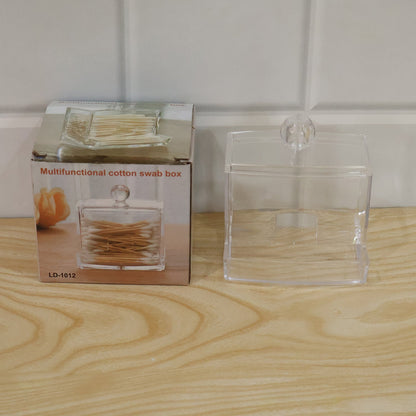 Clear acrylic cotton swab dispenser next to its product packaging box on a wooden surface.