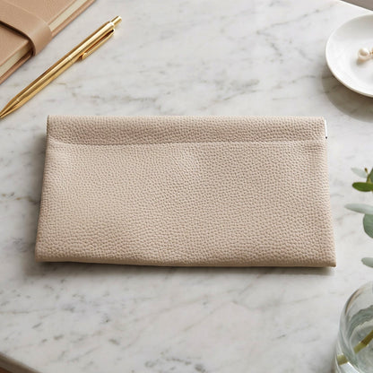 A cream textured faux leather pouch lying flat on a white marble surface, next to a gold pen and a small white dish with