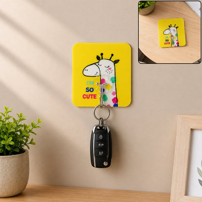 A yellow square wall hook with a cartoon giraffe and text "I'M SO CUTE" holds a black car key on a beige wall.