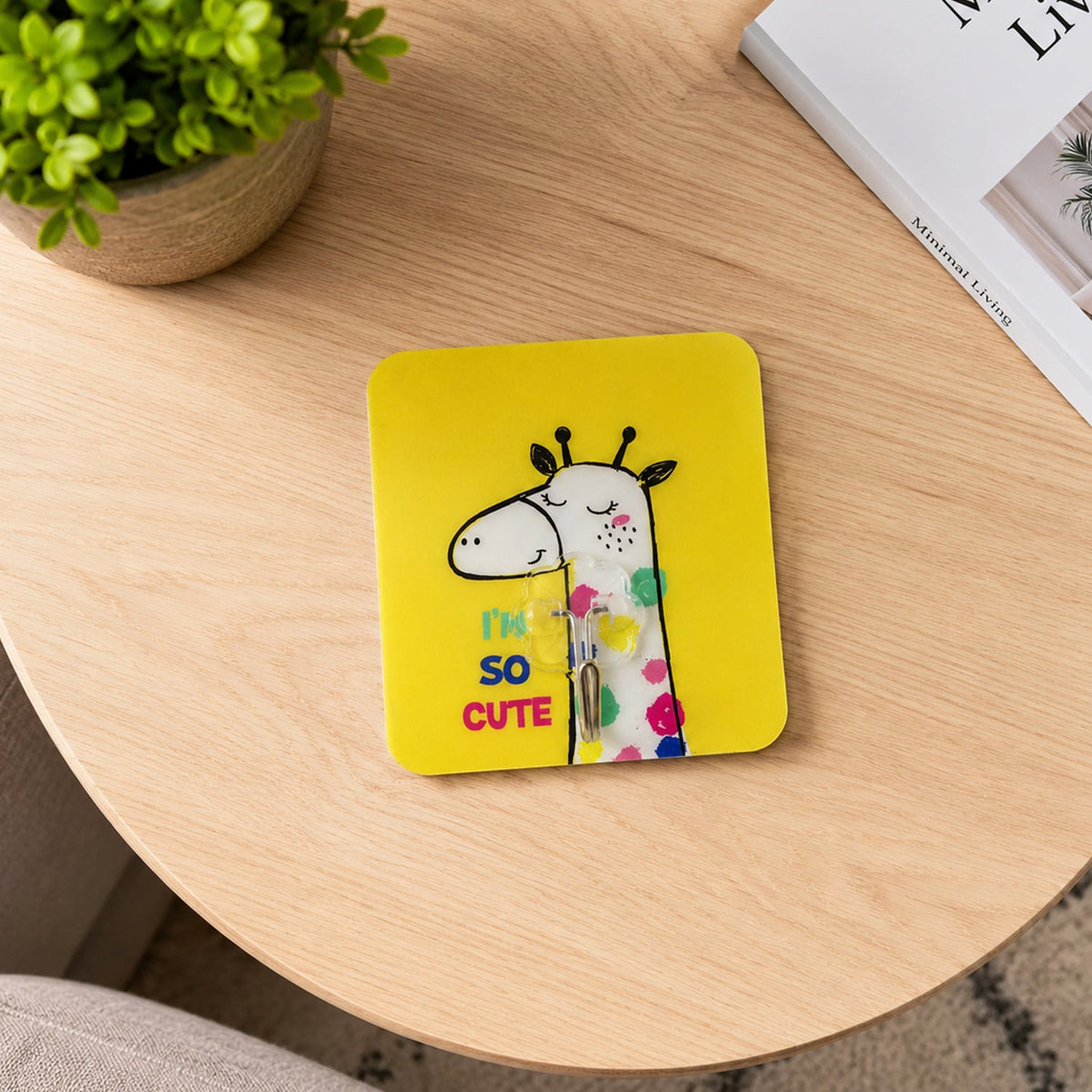 A yellow square wall hook with a cartoon giraffe and text "I'M SO CUTE" rests on a light wooden table next to a green plant.