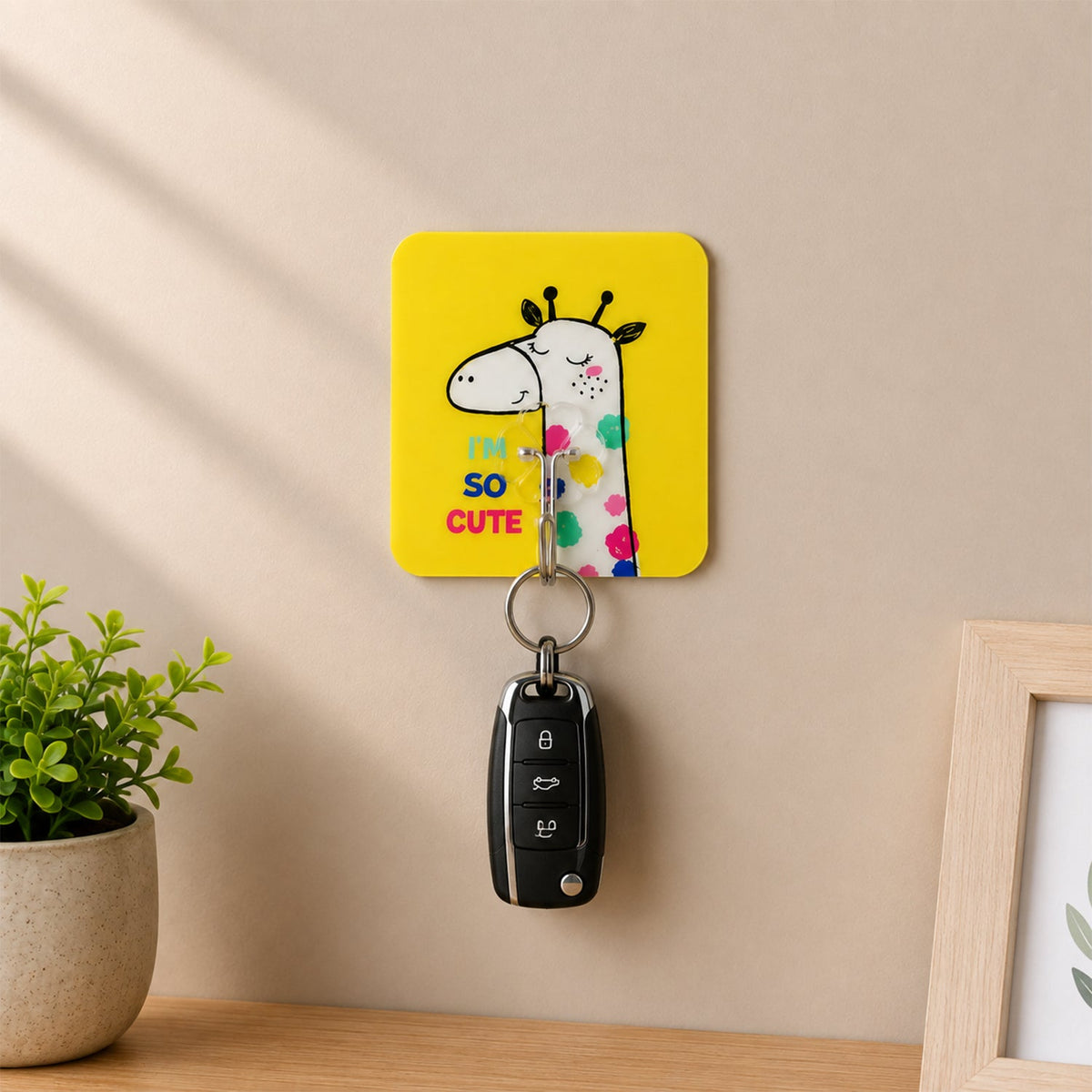 A yellow square wall hook with a cartoon giraffe and text "I'M SO CUTE" holds a black car key, with a potted plant and