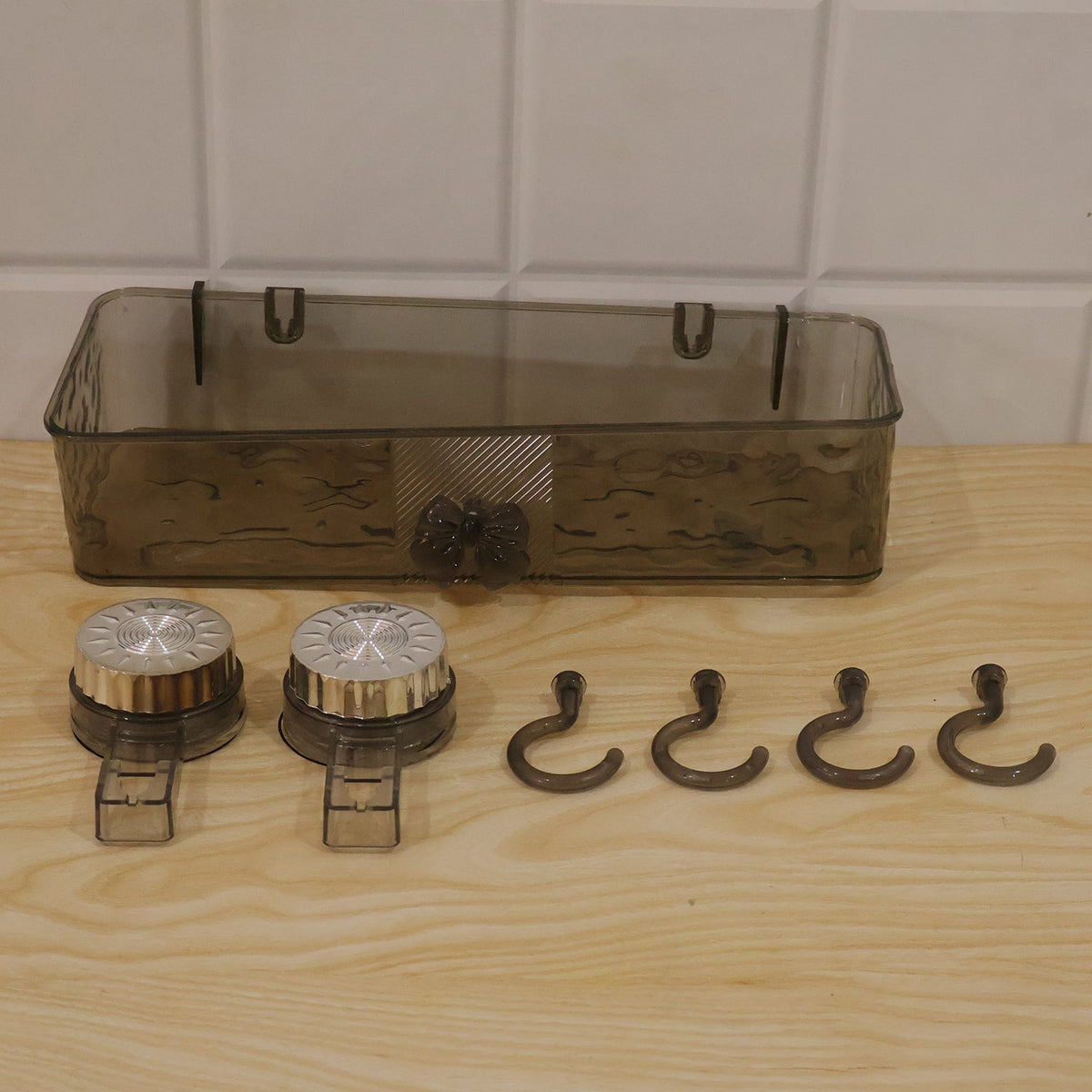 A clear grey plastic caddy, two silver suction cups, and four grey hooks are arranged on a light wooden surface.