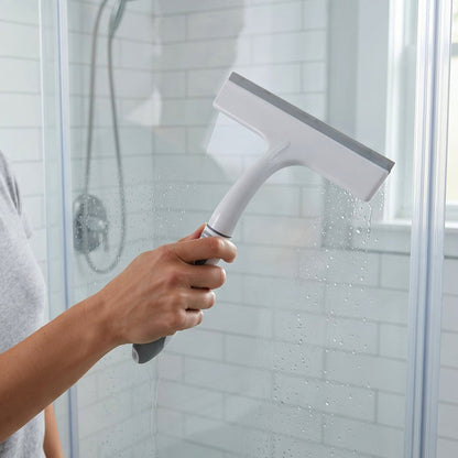A person's hand holding a white and gray glass wiper, cleaning a wet shower door in a bathroom.