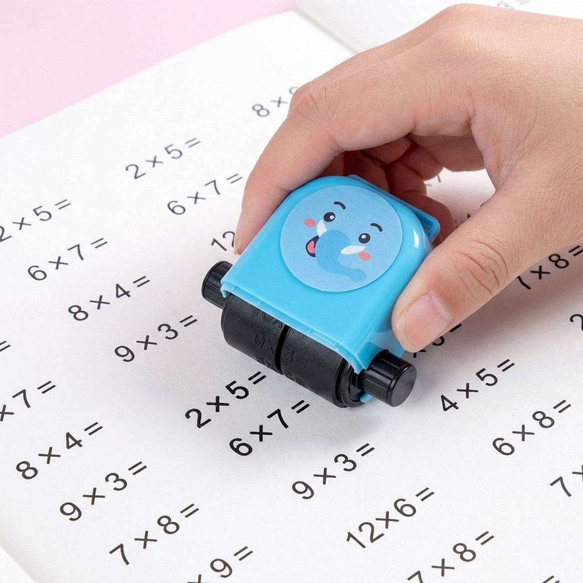 A hand holds a blue elephant-shaped math roller stamp, printing multiplication problems on a white notebook.
