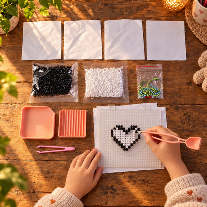Hands carefully arranging black and white fuse beads into a heart shape on a pegboard, surrounded by kit components on a