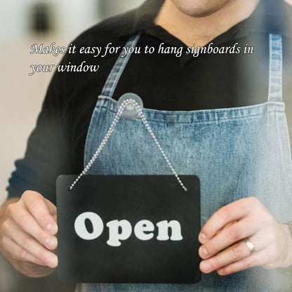 A person in a denim apron holds a black 'Open' sign, attached by a chain to a suction cup hook on a window.