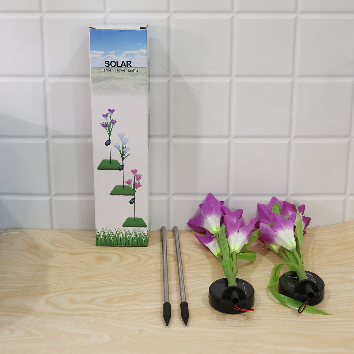 Two disassembled purple solar flower lights, two metal stakes, and a product box are on a wooden surface.
