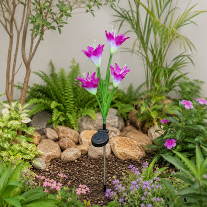 A purple and white solar flower stake light is planted in a landscaped garden bed with rocks and green plants.