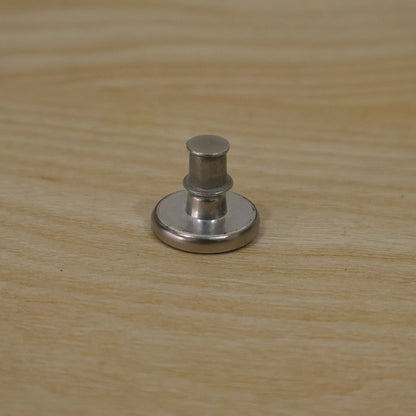 A silver metal button pin with its back lock attached, sitting on a light wooden surface.