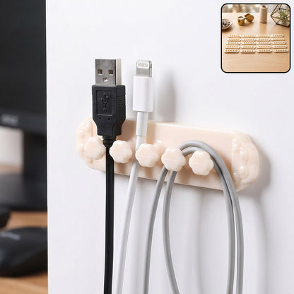 A beige self-adhesive cable organizer clip with five flower-shaped holders, securing black, white, and gray USB cables to a