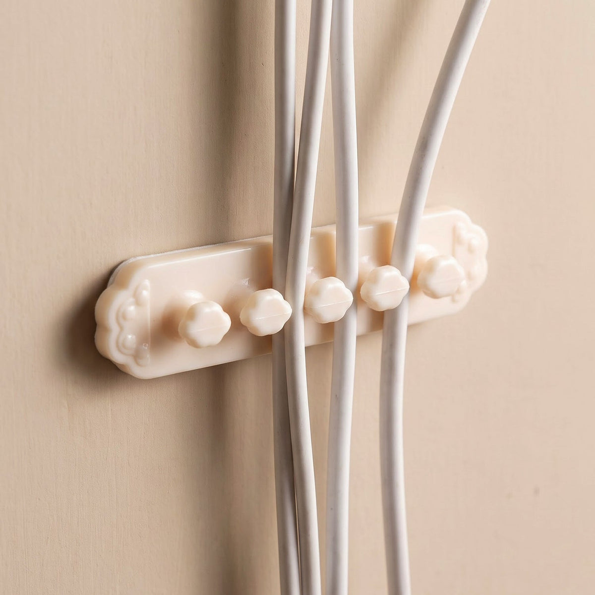 A beige self-adhesive cable organizer clip with five flower-shaped holders securing three white cables on a beige wall.
