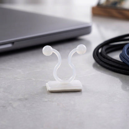A single white plastic self-adhesive plant clip sits on a gray speckled surface, with a laptop and cables blurred in the