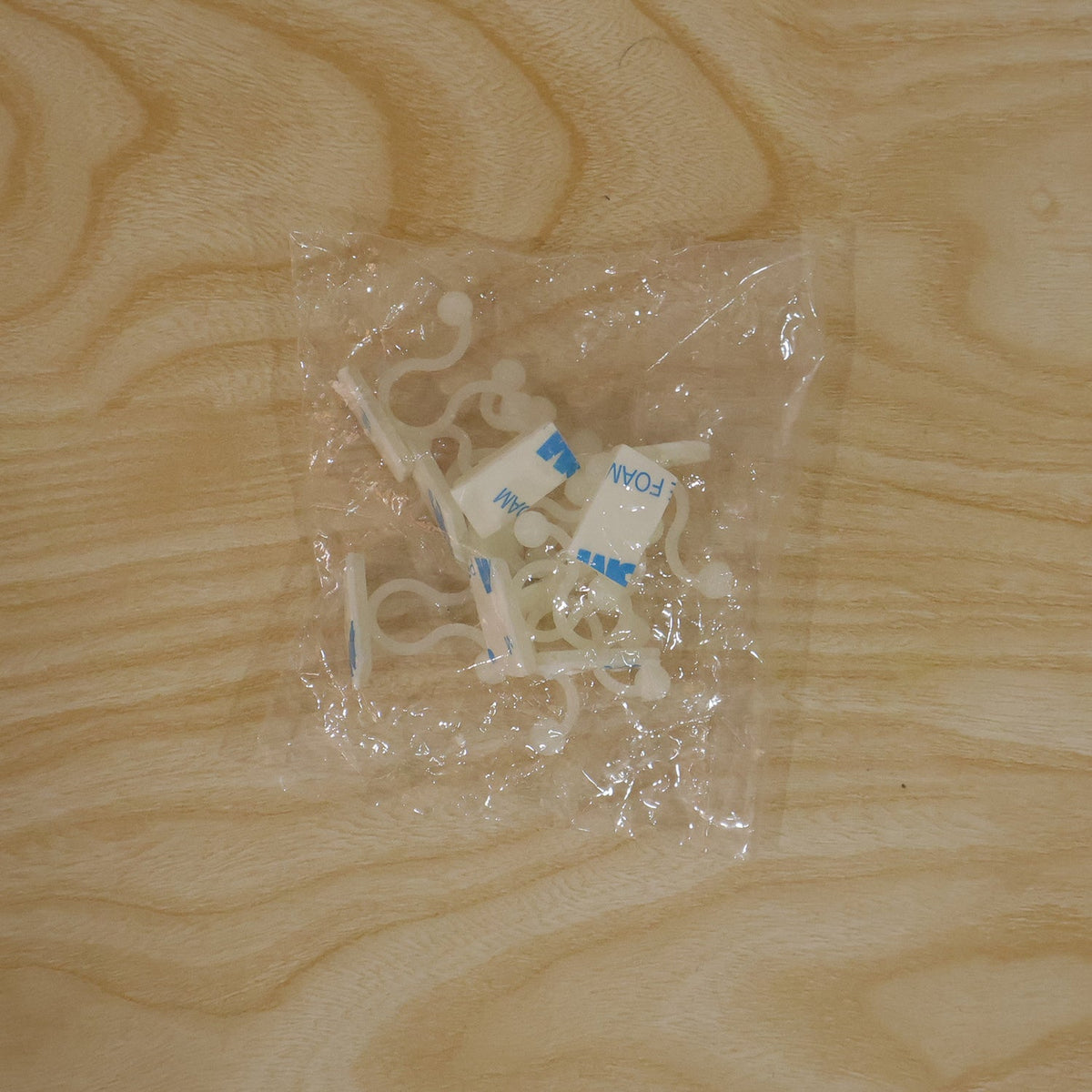Several white plastic self-adhesive plant clips are visible inside a clear plastic bag on a light wooden surface.