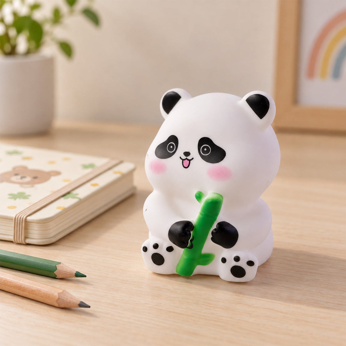 White panda night light with black and pink accents, holding a green bamboo, on a wooden surface with pencils and a notebook.