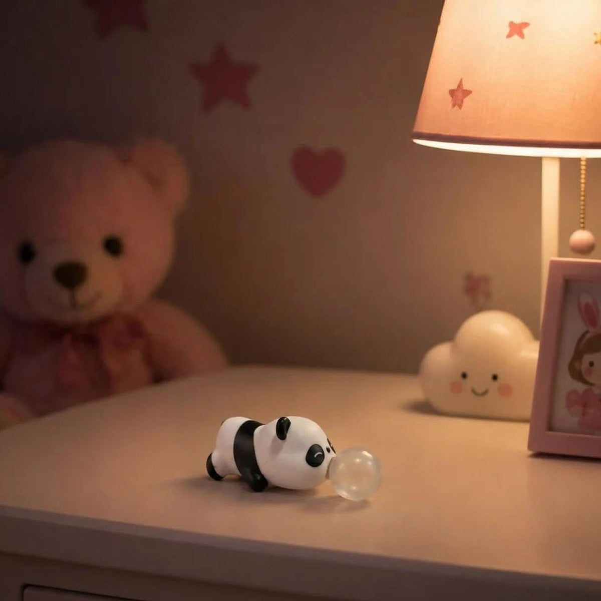 The panda squishy toy with a bubble rests on a bedside table next to a lamp and a teddy bear in a dim room.