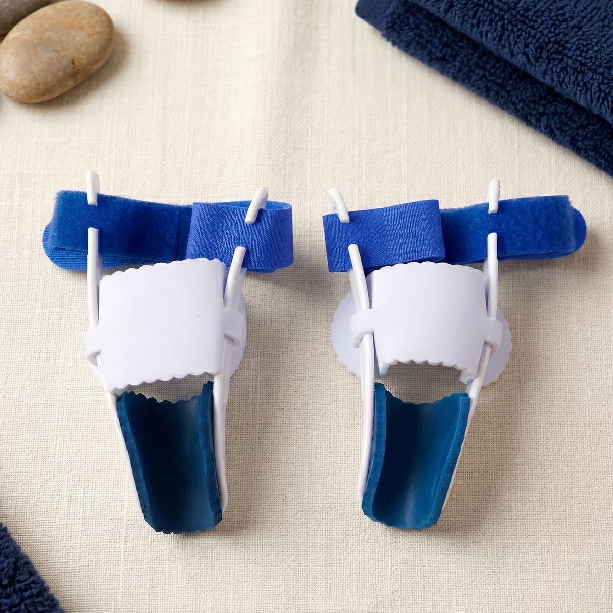 A pair of blue and white toe splints are displayed on a textured surface next to smooth grey stones.