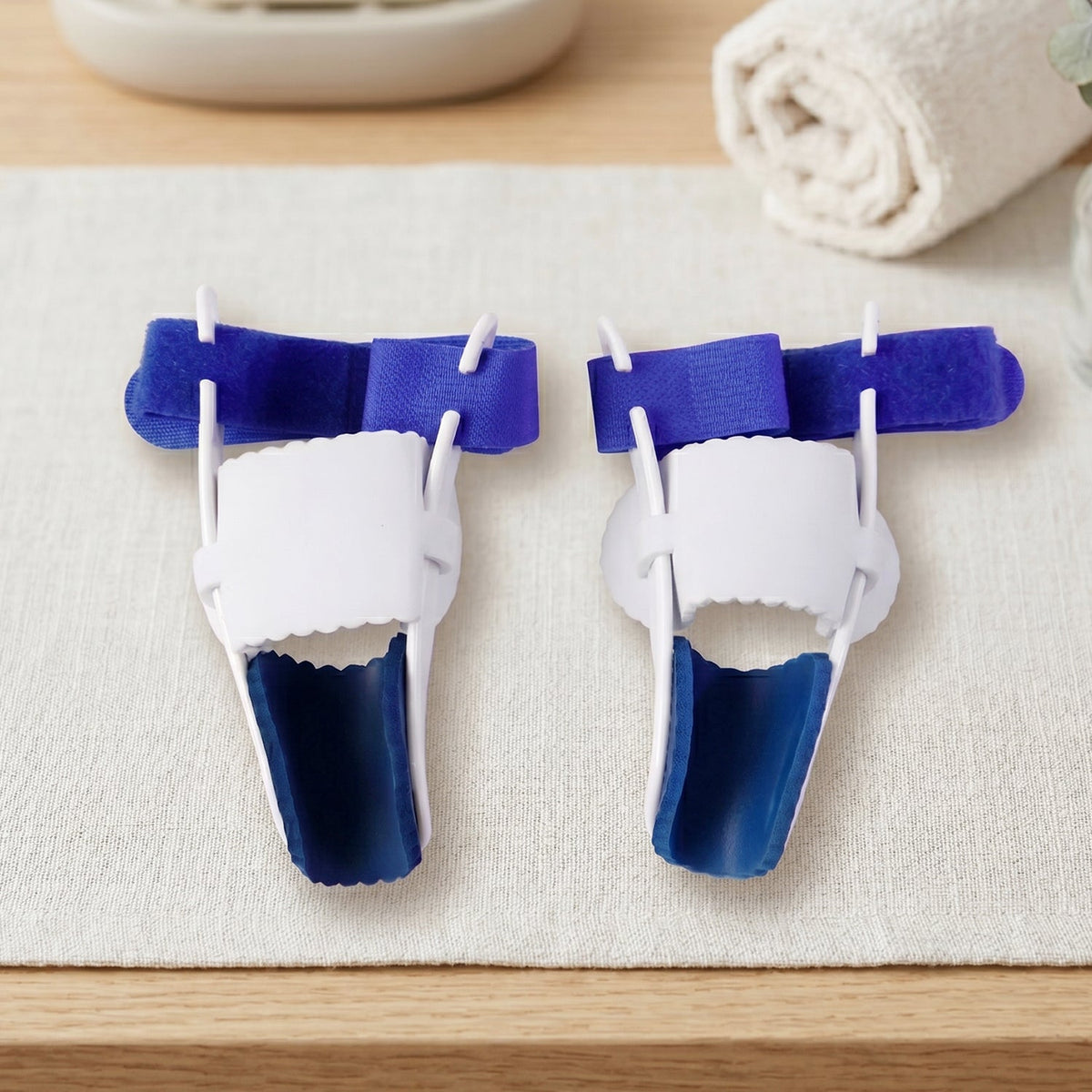 Two blue and white toe splints are neatly arranged on a light fabric surface next to a rolled white towel.