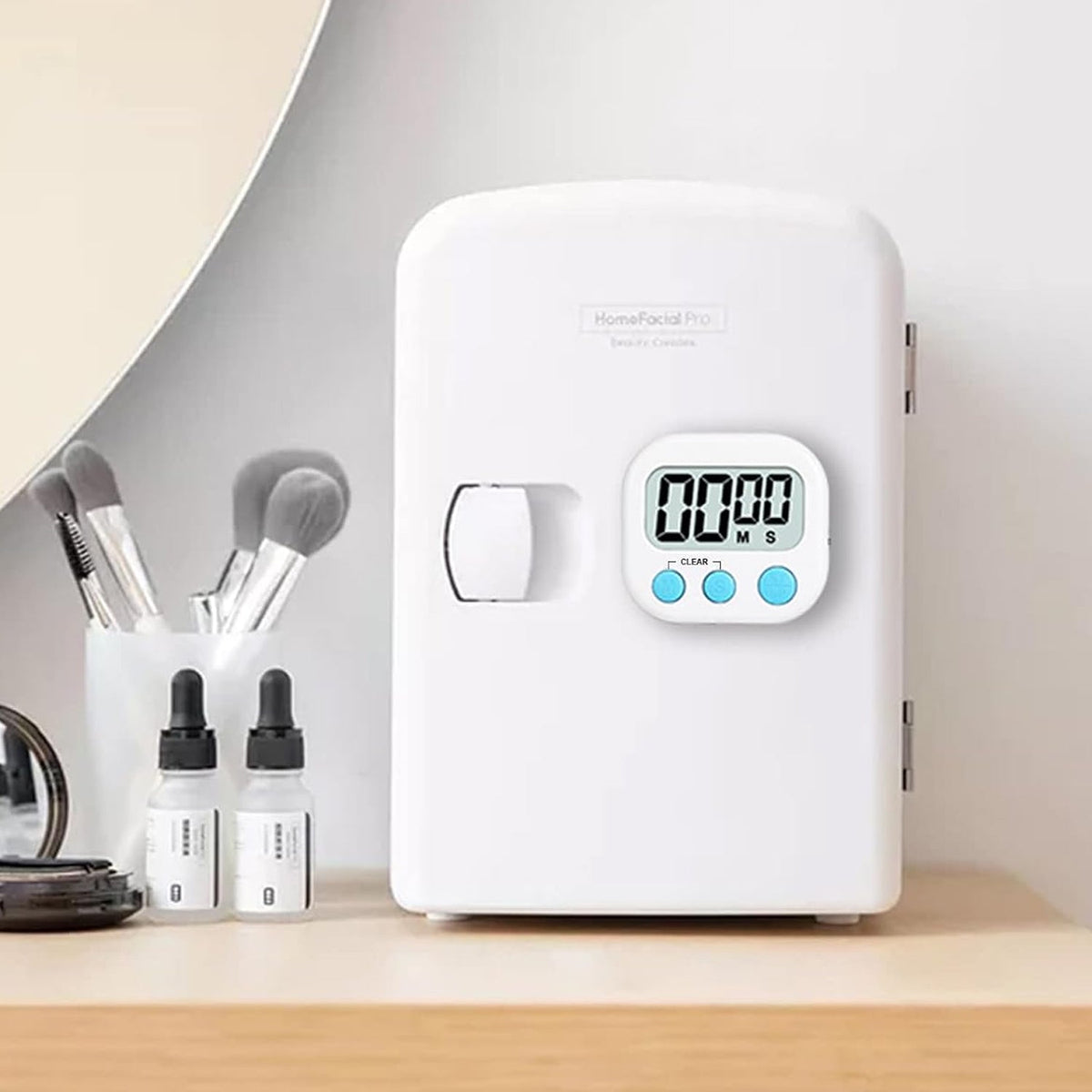 A white digital kitchen timer with blue buttons sits on a wooden desk next to a potted plant and a pen holder.