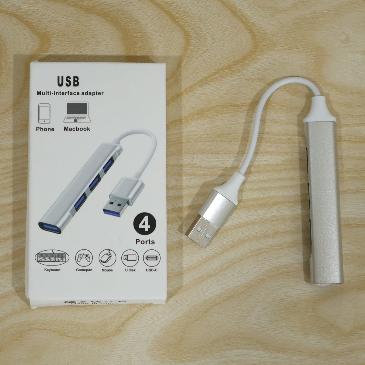 Silver 4-port USB hub next to its white packaging box on a light wooden surface.