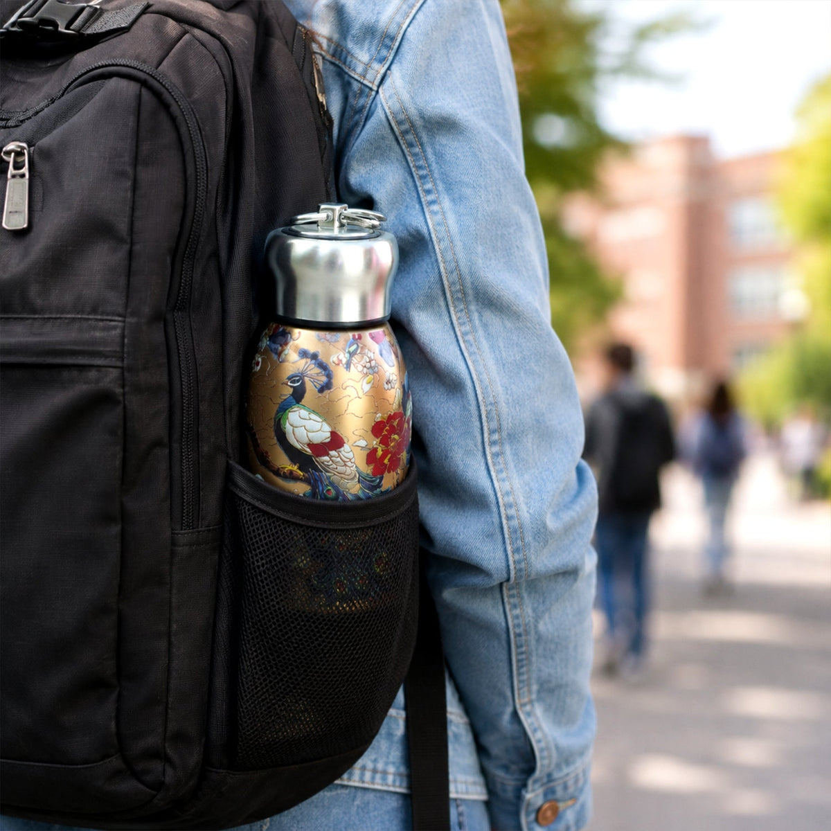Golden stainless steel vacuum bottle with a peacock print tucked into the mesh pocket of a black backpack.