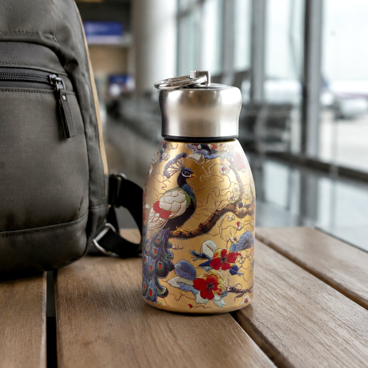 Golden stainless steel vacuum bottle with a peacock and floral print next to a dark grey backpack on a wooden surface.