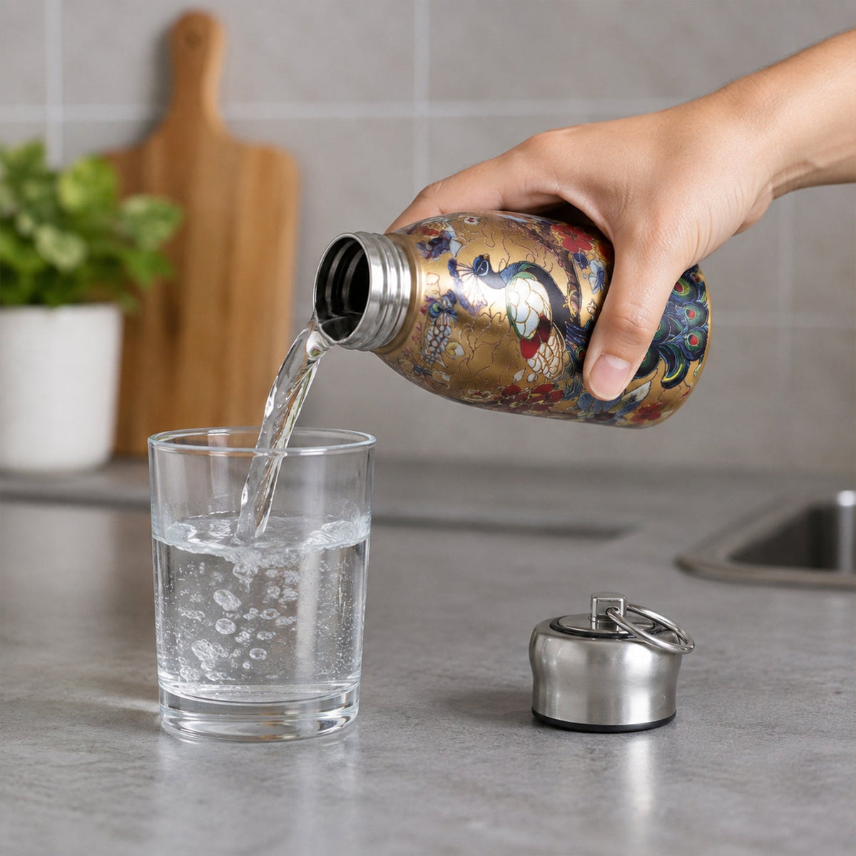 A hand pouring water from the golden stainless steel vacuum bottle with a peacock print into a clear glass.