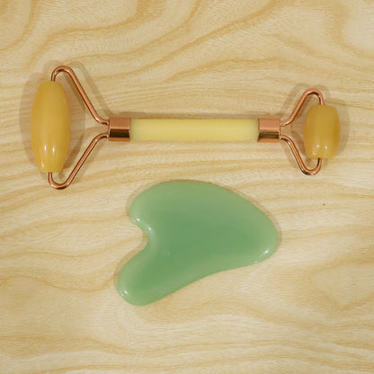 A yellow facial roller with rose gold accents and a green gua sha tool are positioned on a light wooden surface.
