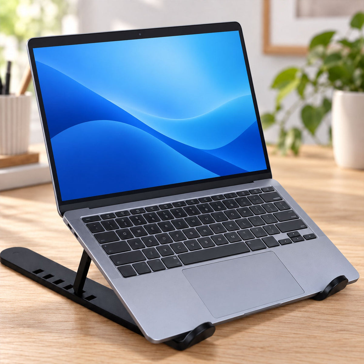 A silver laptop with a blue screen wallpaper sits on a black, low-profile adjustable stand on a light wooden table.