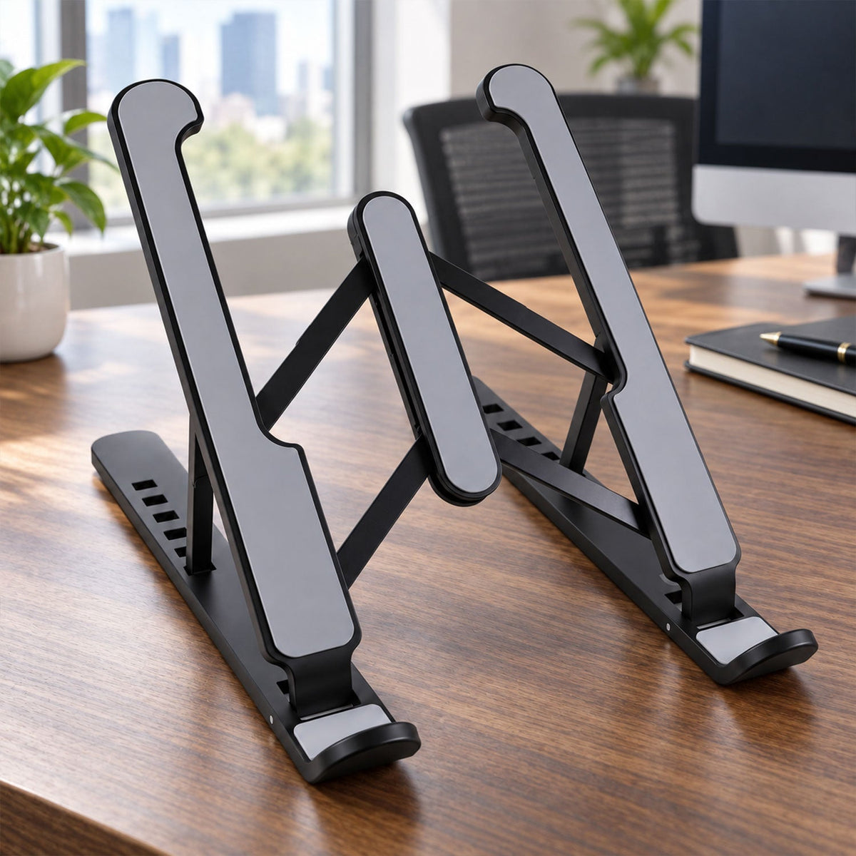 A black and gray adjustable laptop stand is positioned on a wooden desk in a bright office environment.
