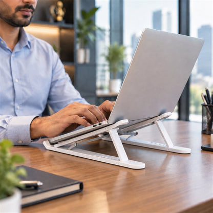 Man working on a silver laptop elevated by a white stand on a wooden desk in a modern office.