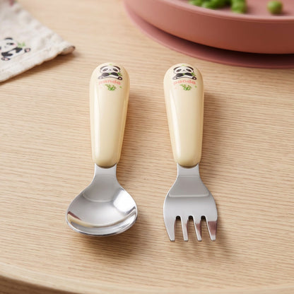 A yellow and silver panda-themed spoon and fork set rests on a wooden surface beside a pink plate.