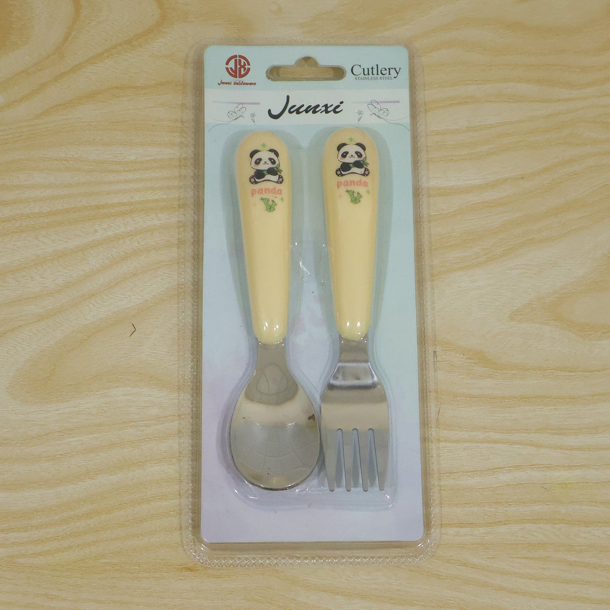 A yellow and silver panda-themed spoon and fork set is sealed in clear plastic packaging on a light wooden surface.