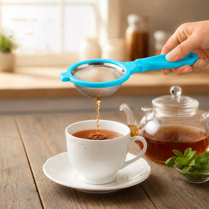A hand holds a blue plastic strainer, pouring dark liquid from a teapot into a white teacup on a wooden table.