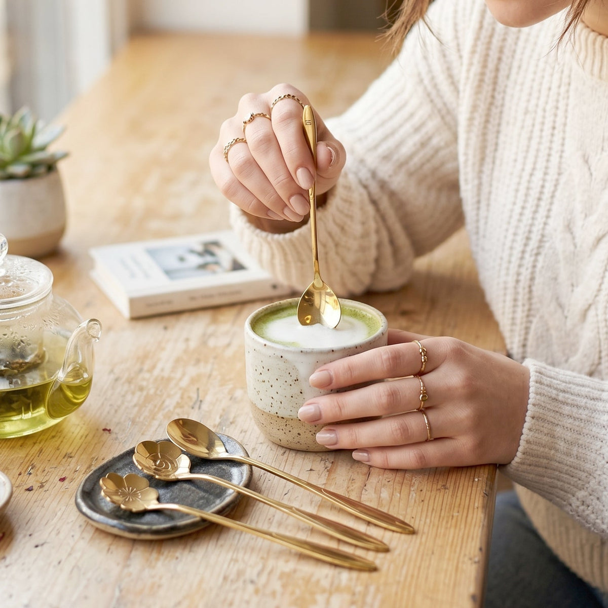 A person stirs a green drink with a gold heart-shaped spoon, three other spoons sit on a small plate.