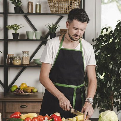 A man with a beard, wearing a white t-shirt and a black apron with green straps, is chopping vegetables on a wooden board in