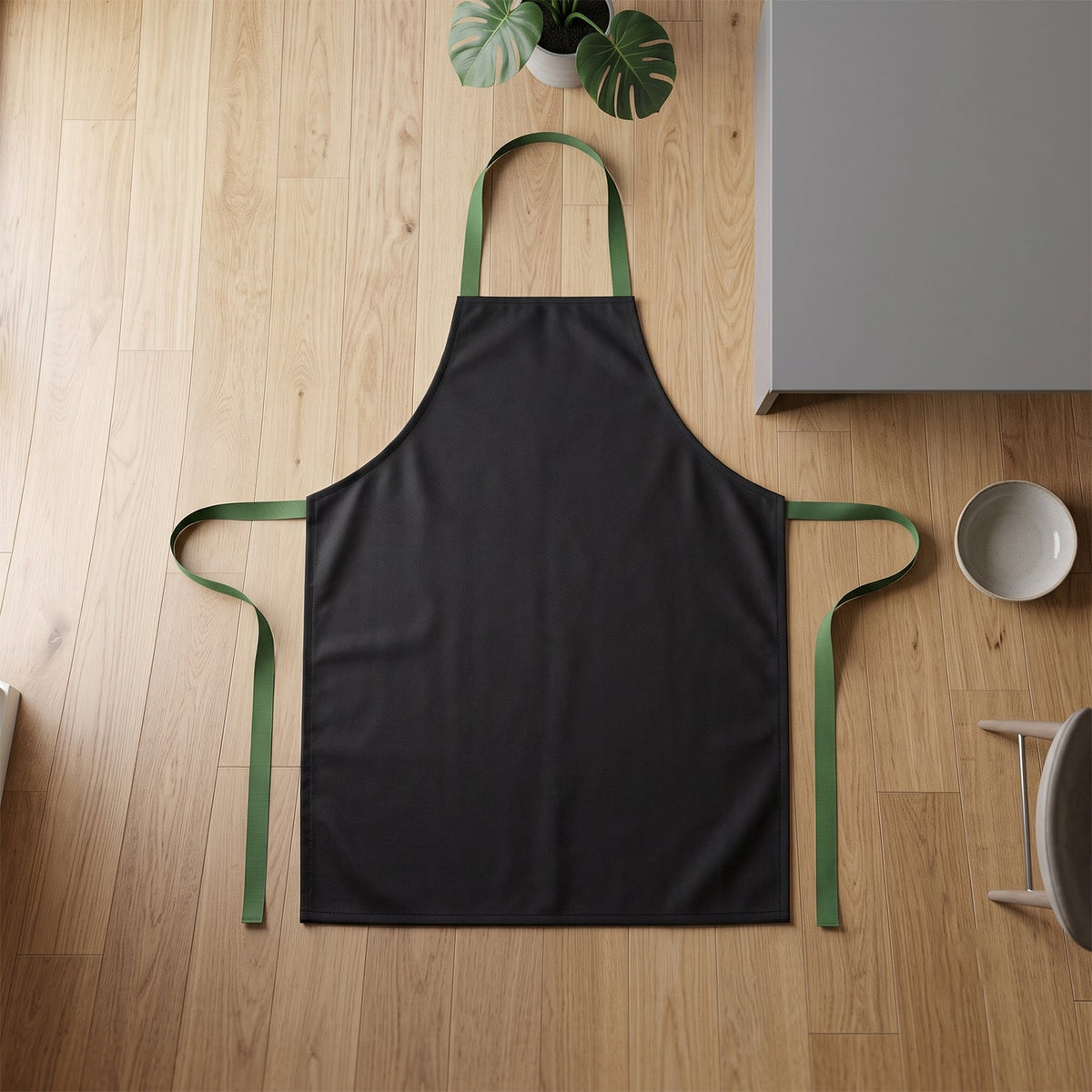 A black apron with green ties laid flat on a light wood floor next to a grey cabinet and plant.