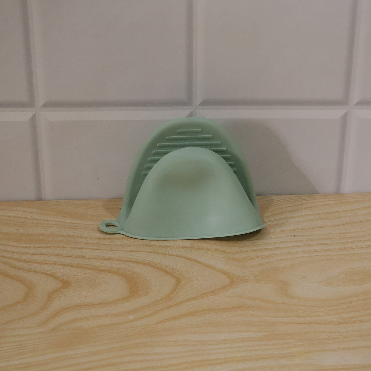 A green silicone mini oven mitt rests on a light wooden surface against a white tiled wall.