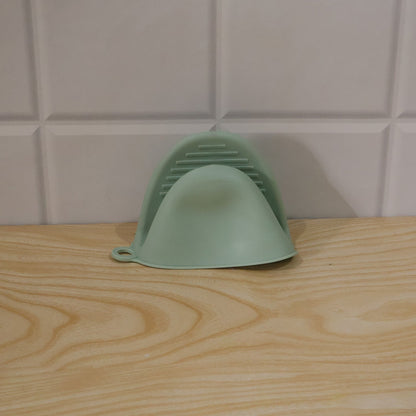 A green silicone mini oven mitt rests on a light wooden surface against a white tiled wall.