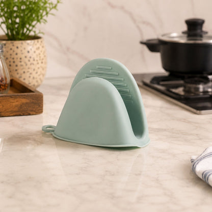A green silicone mini oven mitt sits on a kitchen counter next to a plant and a cooking pot.