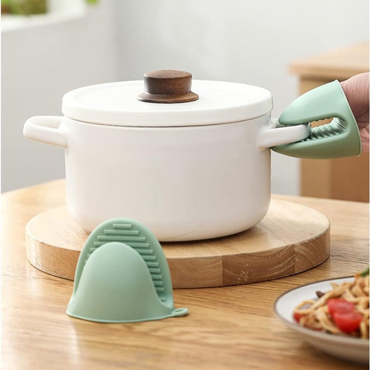 A hand wearing a green silicone mini oven mitt holds the handle of a white lidded pot on a wooden board.
