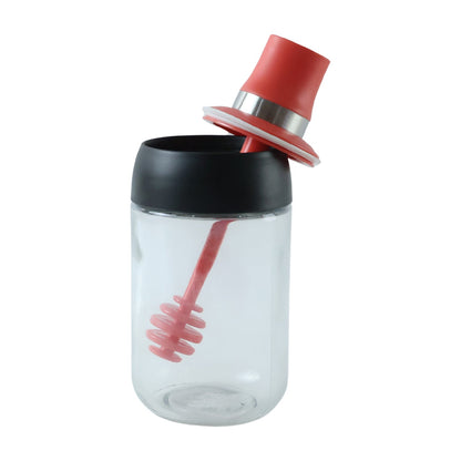 A clear glass dispenser with a red silicone dipper and black lid, with the red cap tilted open.