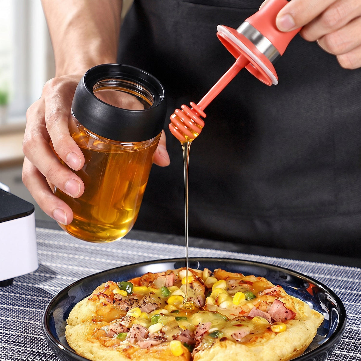 A person drizzles honey from a glass dispenser with a red silicone dipper onto a pizza slice. – view 8