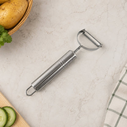 A stainless steel peeler on a light speckled surface next to a basket of potatoes, sliced cucumber, and a green plaid towel.
