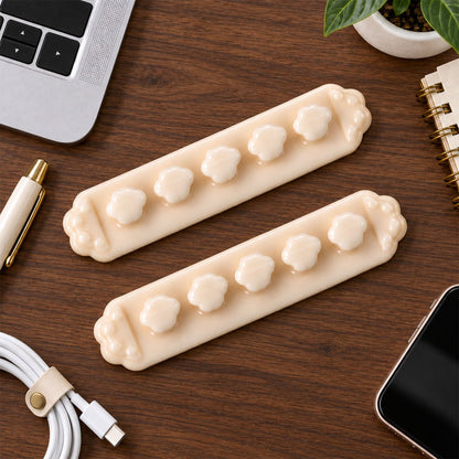 Two beige flower-shaped cable organizer clips on a wooden desk with a laptop, pen, phone, and plant in the background.