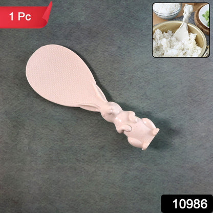A pinkish-beige bunny-handled rice spoon lies on a dark gray surface with a "1 Pc" label and an item number.