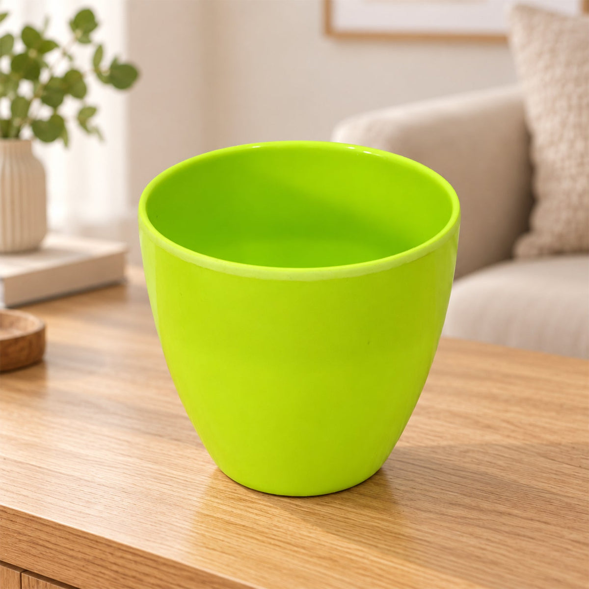 An empty, bright green plastic flower pot with a smooth finish sits on a light wooden table indoors.