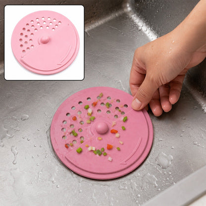 A hand holds a pink silicone drain strainer with food scraps on it, in a stainless steel sink.